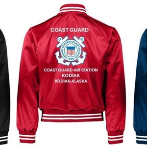 Puede incluir: Tres chaquetas bomber de sat&eacute;n en negro, rojo y azul marino. Cada chaqueta tiene pu&ntilde;os, cuello y dobladillo a rayas blancas y azules. La parte trasera de cada chaqueta muestra las palabras "COAST GUARD" encima de un emblema de la Guardia Costera y el texto "COAST GUARD AIR STATION KODIAK-ALASKA."