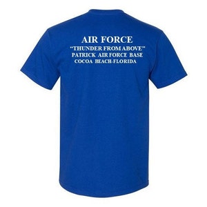 May include: Royal blue t-shirt with white text. The text reads "AIR FORCE", "THUNDER FROM ABOVE", "PATRICK AIR FORCE BASE", and "COCOA BEACH-FLORIDA". The shirt has short sleeves and a crew neck.