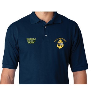 May include: A navy blue polo shirt with yellow embroidered text that reads "USS BIDDLE DLG/CG-34 CRUISER" on the left side and "WORLD'S GREATEST NAVY USN" with an anchor on the right side.