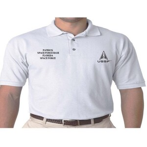 May include: White polo shirt with a black embroidered logo on the right chest. The logo is a stylized triangle with the letters "USSF" inside. The left chest has black embroidered text that reads "Patrick Space Force Base Florida Space Force".