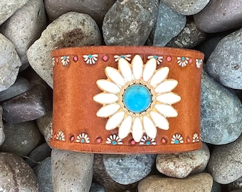 May include: A brown leather cuff bracelet with a white daisy design and a turquoise stone in the center. The bracelet is surrounded by small white and red beads.