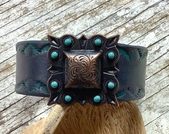 May include: A wide leather cuff bracelet with a square copper-toned metal design featuring a turquoise stone inlay. The bracelet is dark blue with a distressed finish.