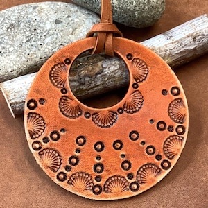 May include: A brown leather pendant with a circular design featuring a repeating pattern of circles and scallops. The pendant is hanging from a brown leather cord.
