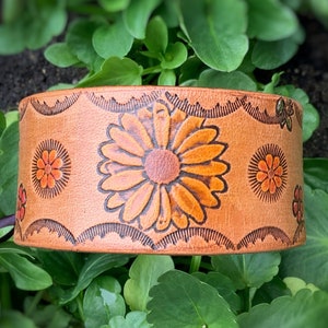 May include: A brown leather cuff bracelet with a floral design. The flowers are orange and yellow and are embossed into the leather. The bracelet is on a green leafy background.