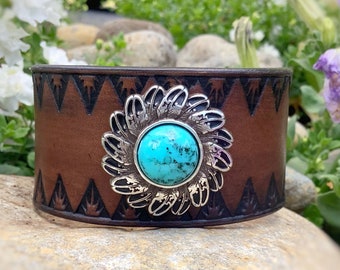 May include: A brown leather cuff bracelet with a silver and turquoise flower design. The bracelet has a geometric pattern on the leather.