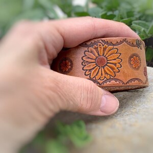 May include: A brown leather bracelet with a floral design. The flowers are orange and brown with green accents.
