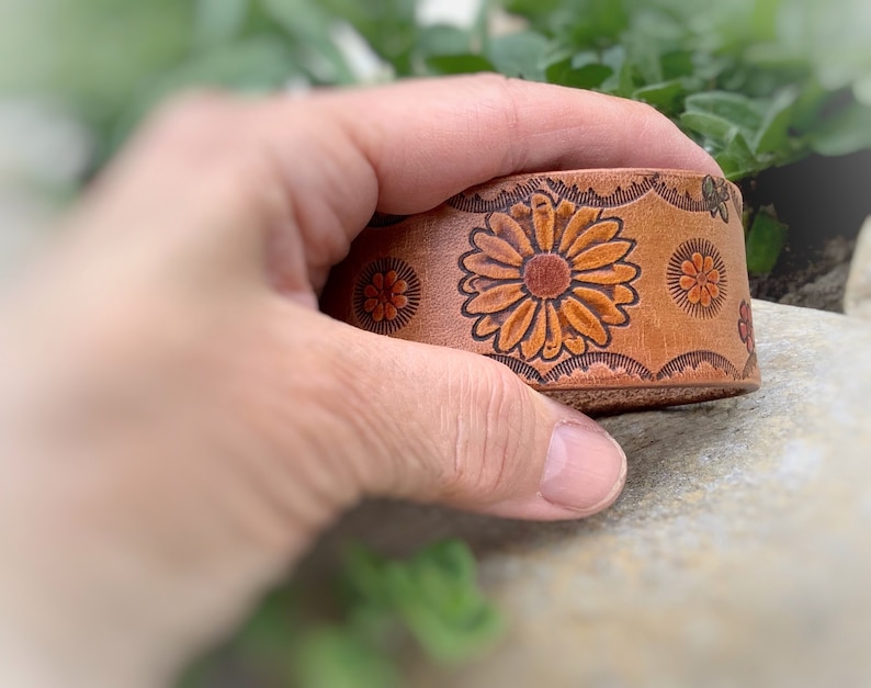 May include: A brown leather bracelet with a floral design. The flowers are orange and brown with green accents.