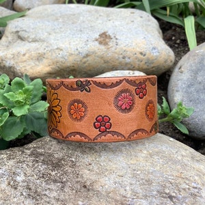 May include: A brown leather cuff bracelet with a floral design. The flowers are in shades of red, orange, and yellow. The bracelet has a scalloped edge.