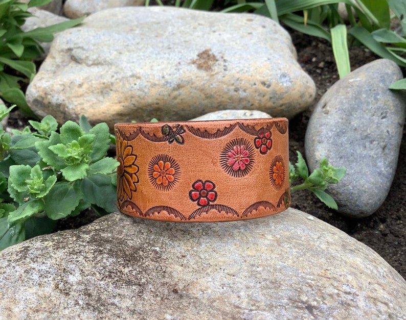 May include: A brown leather cuff bracelet with a floral design. The flowers are in shades of red, orange, and yellow. The bracelet has a scalloped edge.