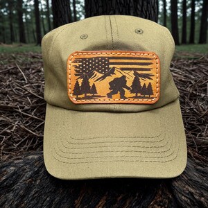 May include: An olive green baseball cap featuring a rectangular leather patch on the front. The patch displays a laser-engraved design of a Bigfoot silhouette, pine trees, and mountain peaks layered over a stylized American flag pattern.