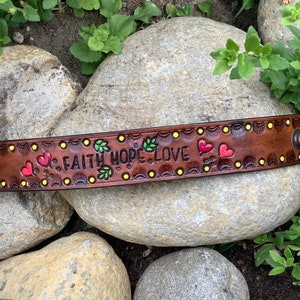 May include: A brown leather bracelet with the words "Faith Hope Love" in yellow paint. The bracelet is decorated with green leaves and red hearts.