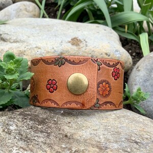 May include: A brown leather bracelet with a floral design and a brass button closure.
