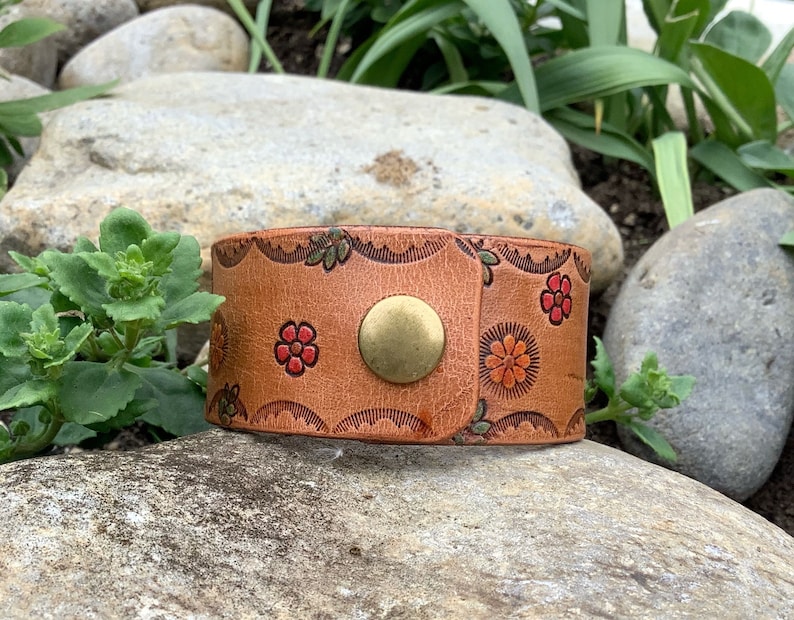 May include: A brown leather bracelet with a floral design and a brass button closure.
