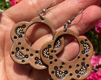 May include: A pair of brown leather earrings with a floral design. The earrings are flower-shaped with a circular centre. The floral design includes white, yellow, and teal accents. The earrings have silver hooks.