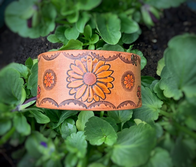 May include: A brown leather cuff bracelet with a floral design. The flowers are orange and yellow and are embossed into the leather. The bracelet is on a green leafy background.