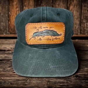 May include: A dark green baseball cap featuring a rectangular tan leather patch stitched to the front. The patch displays a hand-drawn illustration of a single feather and the text "He will cover you with His feathers" in black script.