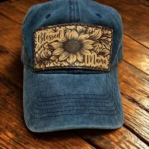 May include: A blue denim baseball cap featuring a rectangular leather patch centered on the front. The patch is laser-engraved with a large sunflower illustration and the text Blessed Mama in a cursive script font, with visible stitching.