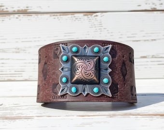 May include: A wide brown leather cuff bracelet featuring an embossed floral pattern and a large square metal centerpiece. The centerpiece has a copper-toned, swirling engraved square surrounded by a silver-toned frame with eight small turquoise beads.