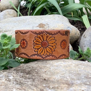 May include: A brown leather bracelet with a floral design. The flowers are orange, yellow, and brown. The bracelet is on a rock.