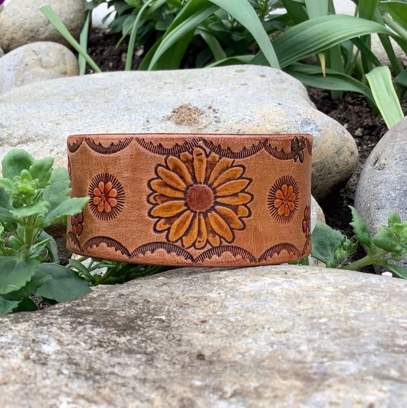 May include: A brown leather bracelet with a floral design. The flowers are orange, yellow, and brown. The bracelet is on a rock.