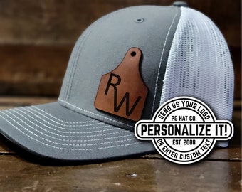 May include: Gray and white trucker hat with a leather patch featuring the letters "RW". The hat has a curved brim and a mesh back. The text "PERSONALIZE IT!" is also visible.
