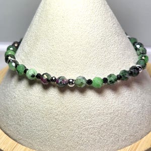 May include: A beaded bracelet featuring green and ruby-colored gemstones, interspersed with small black beads and silver-colored beads. The bracelet is displayed on a white, cone-shaped stand with a wooden base.