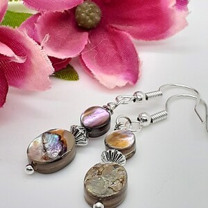 May include: A pair of silver dangle earrings with iridescent brown and purple shell beads. The earrings have a silver hook closure.