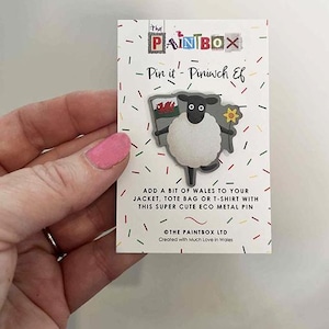 May include: A metal pin featuring a cartoon sheep holding a Welsh flag and a flower. The pin is on a white card with the text "Pin it - Piniwch Ef" and "The Paintbox" logo. The card has colorful confetti.