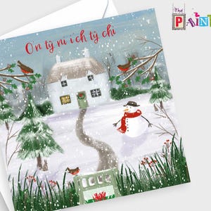 May include: A Christmas card featuring a winter scene with a white cottage, snow, and a snowman wearing a hat and scarf. The card has text in a foreign language and is decorated with birds and evergreen trees. The card is in an envelope.