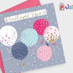 Puede incluir: Una tarjeta gris con el texto "Penblwydd Hapus" en letras doradas. La tarjeta presenta un diseño de globos coloridos con lunares, rayas y confeti. Los globos están atados con cuerdas de cuentas.