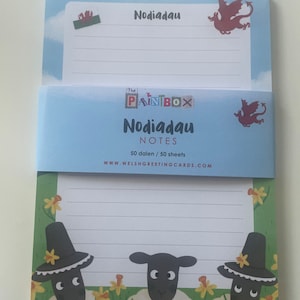 May include: A blue and white notepad with a Welsh dragon design. The notepad says "Nodiadau Notes" and "50 dalen / 50 sheets". The back of the notepad has a design of three sheep with black hats in a field of yellow daffodils.