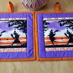 May include: Two purple and orange Halloween pot holders with a silhouette of two witches flying with broomsticks in front of a purple and orange sunset. The pot holders have the word "Halloween" printed on them.