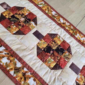 以下が含まれることがあります： オレンジ、赤、茶色、黄色の色合いで様式化されたカボチャの繰り返し模様が特徴のキルトテーブルランナー。カボチャはクリーム色の背景に配置され、秋の葉のボーダーが付いています。