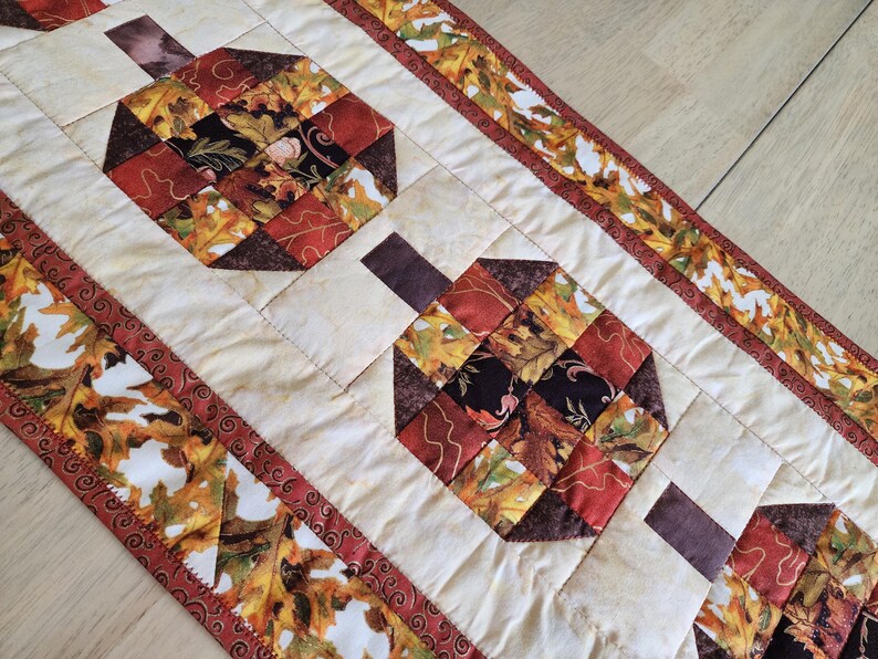 以下が含まれることがあります： オレンジ、赤、茶色、黄色の色合いで様式化されたカボチャの繰り返し模様が特徴のキルトテーブルランナー。カボチャはクリーム色の背景に配置され、秋の葉のボーダーが付いています。