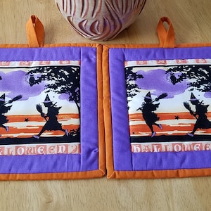 May include: Two purple and orange Halloween pot holders with a vintage design of two witches flying on broomsticks with the word "Halloween" printed on the bottom.