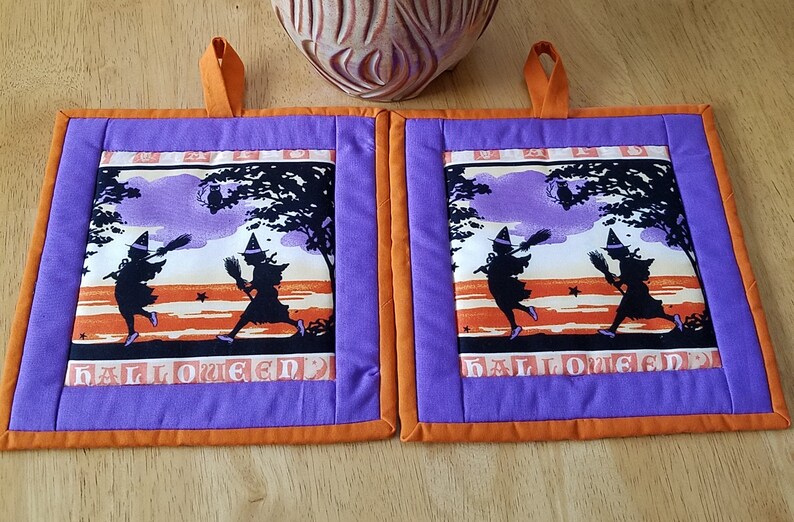 May include: Two purple and orange Halloween pot holders with a vintage design of two witches flying on broomsticks with the word "Halloween" printed on the bottom.