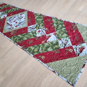 May include: Quilted table runner with a festive design. Features a central diamond pattern with birds and floral motifs on a light blue background. Alternating red and green sections with intricate quilting, creating a seasonal, decorative piece.