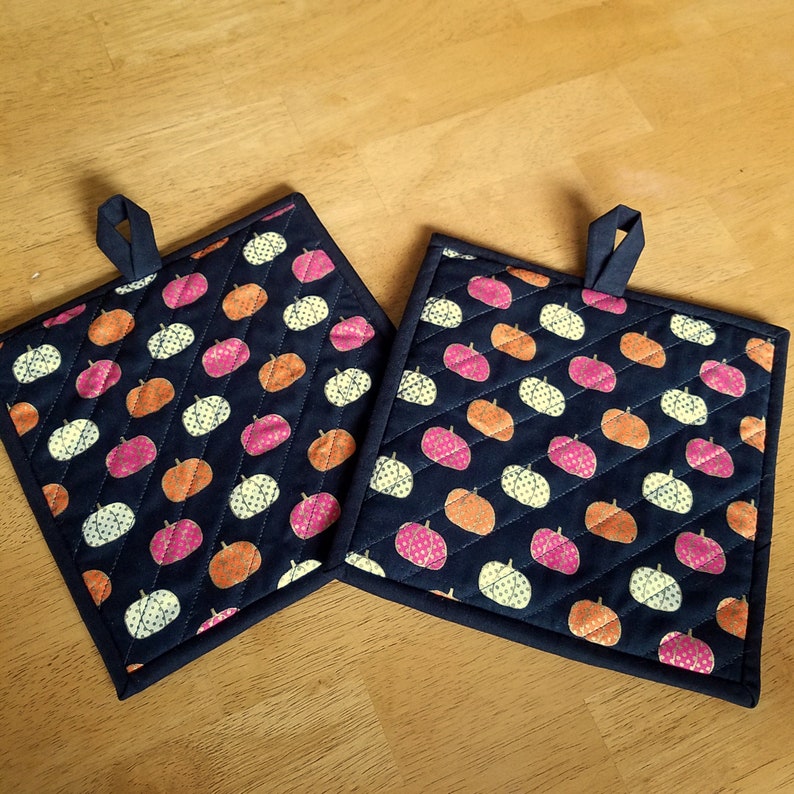 May include: Two dark blue quilted pot holders with a white, orange, and pink pumpkin pattern.