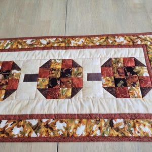 以下が含まれることがあります： 秋をテーマにしたキルトテーブルランナー。 ランナーは、秋の色合いのパッチワークデザインの3つの八角形のパッチを備えています。 境界線とパッチには、オレンジ、黄色、茶色の葉のパターンがあります。