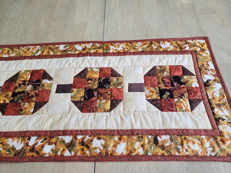 以下が含まれることがあります： 秋をテーマにしたキルトテーブルランナー。 ランナーは、秋の色合いのパッチワークデザインの3つの八角形のパッチを備えています。 境界線とパッチには、オレンジ、黄色、茶色の葉のパターンがあります。