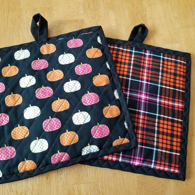 May include: Two black quilted pot holders. One has a black background with white and pink pumpkins. The other has a black, orange, and pink plaid pattern.