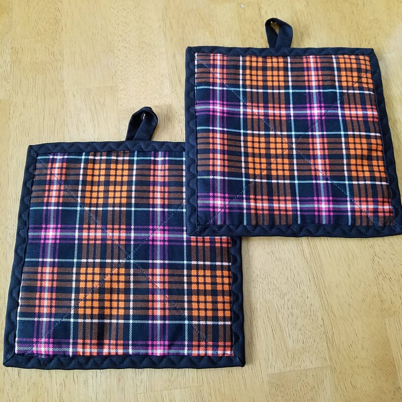 May include: Two black and orange plaid pot holders with a black border. The pot holders are quilted and have a loop for hanging.