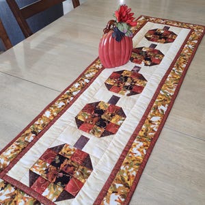 以下が含まれることがあります： 秋をテーマにしたキルトのテーブルランナー。 ランナーは、4つのカボチャ型のデザインが施されたクリーム色のセンターを備えています。 カボチャは、さまざまな秋色の生地で作られています。 境界線には、バーガンディのトリムが付いた葉のパターンがあります。 花瓶と花がランナーの上にあります。