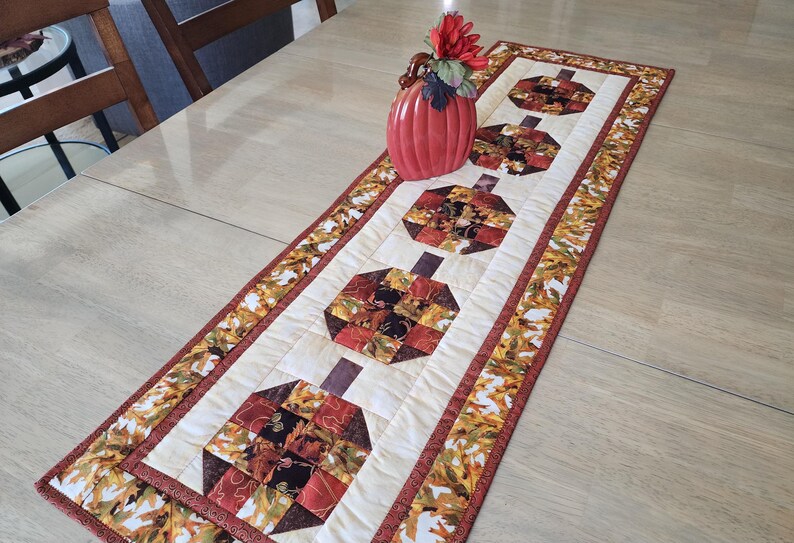 以下が含まれることがあります： 秋をテーマにしたキルトのテーブルランナー。 ランナーは、4つのカボチャ型のデザインが施されたクリーム色のセンターを備えています。 カボチャは、さまざまな秋色の生地で作られています。 境界線には、バーガンディのトリムが付いた葉のパターンがあります。 花瓶と花がランナーの上にあります。