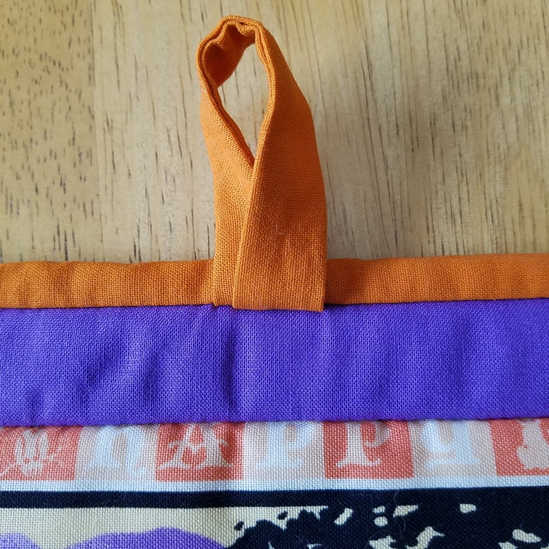 May include: A purple and orange fabric tote bag with a loop handle. The bag has a patterned fabric with a white background and orange and black designs.