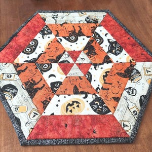 May include: Hexagon-shaped Halloween-themed quilted placemat. The placemat features orange, black, and white patterned fabric with cat, pumpkin, and bottle designs. The border is a dark gray fabric.