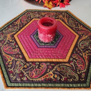 May include: A quilted, hexagonal table runner with a paisley design in shades of burgundy, pink, gold, and green. A red candle sits in the center of the runner, which measures approximately 20 inches across.