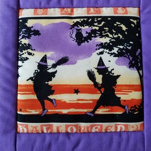 May include: A purple fabric square with a white fabric square inside. The white fabric square has a Halloween scene with two witches flying on broomsticks, an owl in a tree, and the words "Halloween" at the bottom.