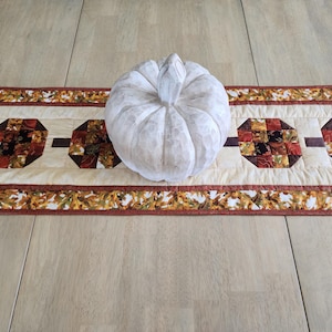 以下が含まれることがあります： 白く質感のあるカボチャが、秋をテーマにしたテーブルランナーの上に置かれています。ランナーは、茶色、赤、オレンジの色合いの幾何学模様が施されたクリーム色のセンターを備えています。縁は秋の葉で飾られています。