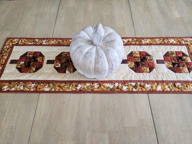 以下が含まれることがあります： 白く質感のあるカボチャが、秋をテーマにしたテーブルランナーの上に置かれています。ランナーは、茶色、赤、オレンジの色合いの幾何学模様が施されたクリーム色のセンターを備えています。縁は秋の葉で飾られています。